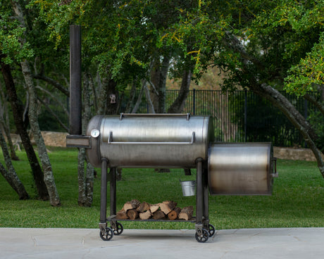 A large silver-colored American-made steel barbecue pit with a removable smokestack, set on a grassy area with trees in the background.